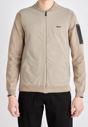 Mann trägt beige Reißverschlussjacke mit gestrickten Ärmeln und "BOSS"-Logo auf der Brust, kombiniert mit schwarzer Hose, steht vor weißem Hintergrund.