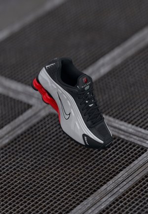 Black and gray athletic shoe with red cushioned sole, black laces, and swoosh logo resting on metal grid surface.
