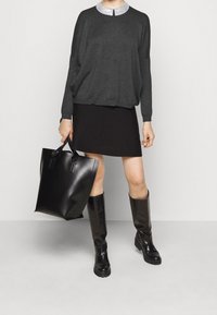 Kobieta w ciemnoszarym swetrze z srebrnym kołnierzykiem, czarnej spódnicy, czarnych botkach do kolan, trzymająca dużą czarną skórzaną torbę.