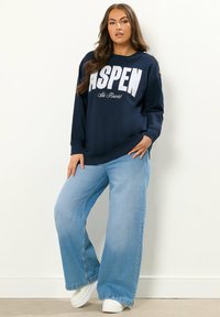 Felpa blu navy con la scritta "ASPEN Ski Resort" in bianco, abbinata a jeans chiari a gamba larga e sneakers bianche. Tessuto dalla consistenza liscia.