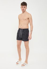 Pantaloni da nuoto neri con cordoncino, caratterizzati da accenti laterali rosa e blu. Indossati con sandali bianchi, il modello è senza maglietta e ha i capelli corti.