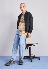Giacca leggera nera sopra una camicia a camouflage marrone e verde, abbinata a jeans azzurri chiari e sneakers nere. Sgabello in legno sullo sfondo.
