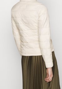 Lyse beige puffjakke med vandrette quiltede paneler, der har en høj krave og skjulte sidelommer, parret med en mørkegrøn plisseret nederdel.