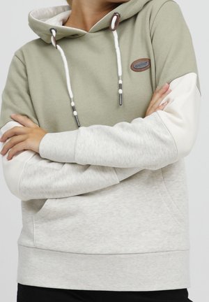 Personne portant un sweat à capuche bicolore vert et gris clair avec cordons blancs, bras croisés, debout devant un fond uni.