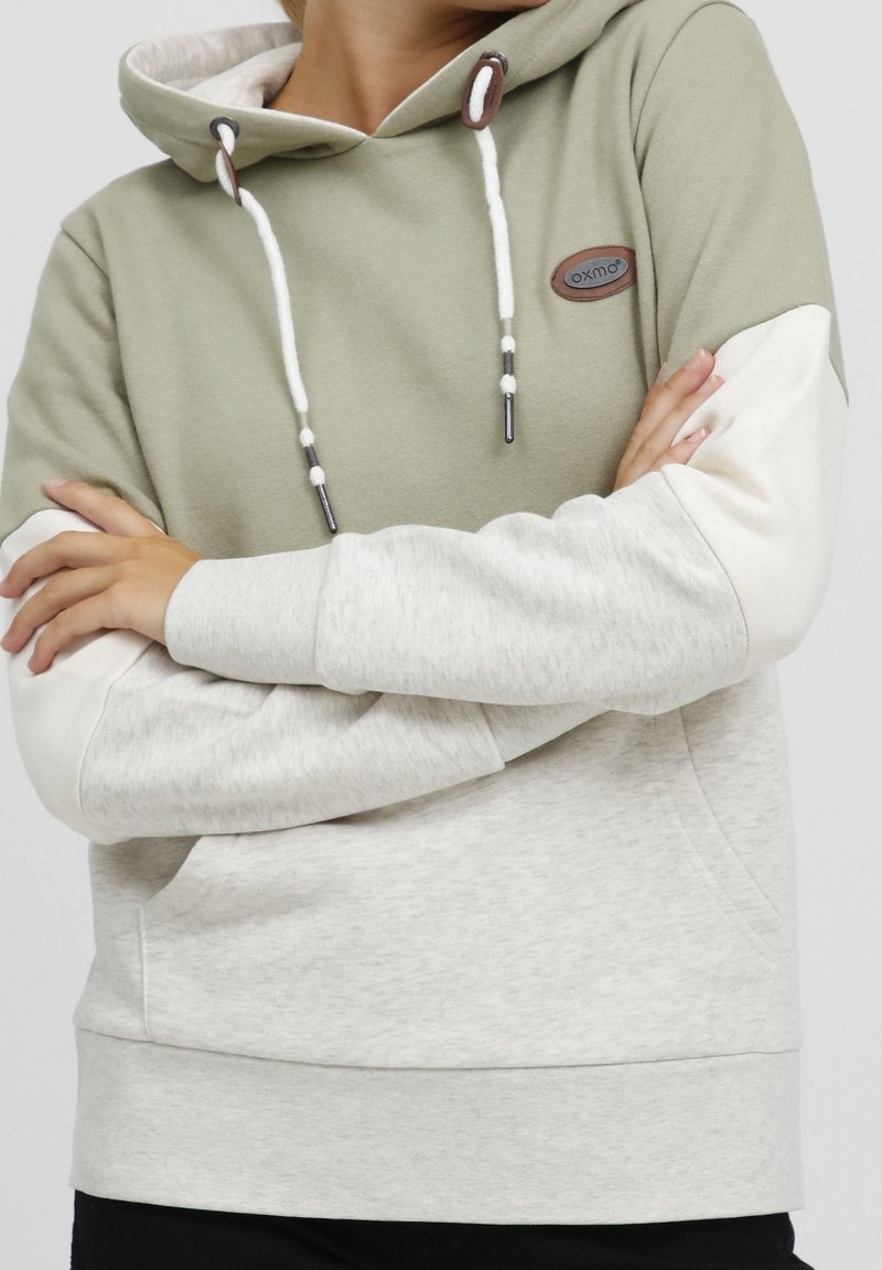 Personne portant un sweat à capuche bicolore vert et gris clair avec cordons blancs, bras croisés, debout devant un fond uni.