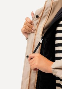 Beige Jacke mit Reißverschluss vorne, zweifarbigen Stoff, sichtbaren Druckknöpfen und einer glatten Textur. Das Innenfutter ist schwarz mit einem gestreiften Detail.