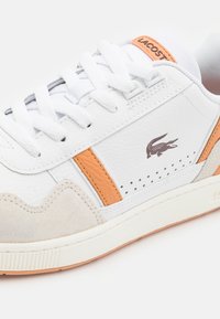 Basket blanche avec des accents orange, logo Lacoste sur le côté, lacets blancs et bout en daim beige, présentée sur fond uni.