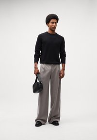 Pulover negru din tricot, pantaloni plisati gri și o mică geantă din țesătură neagră. Pantofi negri completează ținuta. Fundal neutru.