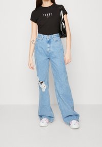 Zwarte t-shirt met korte mouwen met "TOMMY" logo, gecombineerd met lichtblauwe hoge taille spijkerbroek met een gescheurde knie. Witte sneakers en een zwarte handtas.