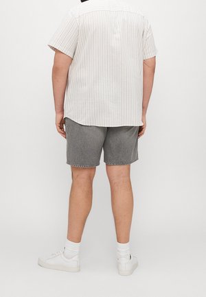 Mann mit weißem kurzärmligem gestreiftem Hemd, grauen Jeans-Shorts, weißen Socken und weißen Sneakers, steht vor einem einfarbigen Hintergrund.