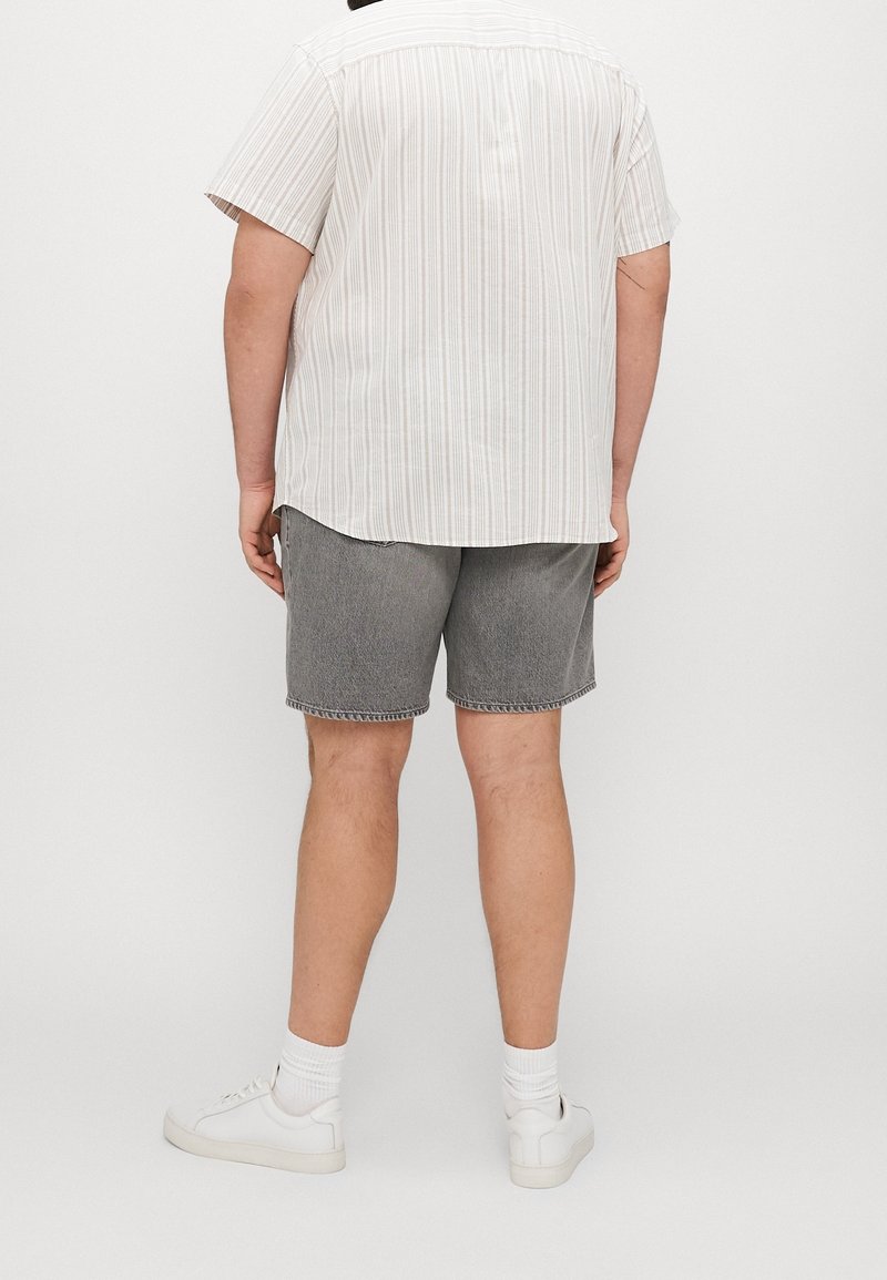 Homme portant une chemise blanche à manches courtes rayée, un short en jean gris, des chaussettes blanches et des baskets blanches, debout devant un fond uni.