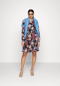 Vestido floral com fundo preto, apresentando flores rosas e azuis, combinado com um suéter sem mangas azul claro e sandálias de salto transparente.
