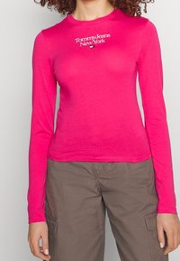 Osoba v jasně růžovém tričku s dlouhým rukávem a logem "Tommy Jeans New York" a tmavě hnědými cargo kalhotami, stojící na jednobarevném pozadí.