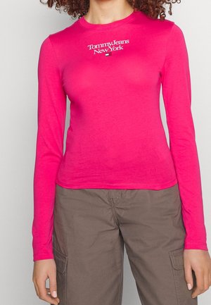Persoon in een felroze lange mouw shirt met het logo "Tommy Jeans New York" en donkerbruine cargo-broek, staande tegen een effen achtergrond.