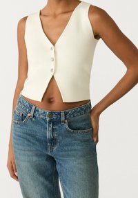 Kvinna i en ärmlös krämfärgad knäppt crop top och högmidjade blå denimjeans med en hand på höften.