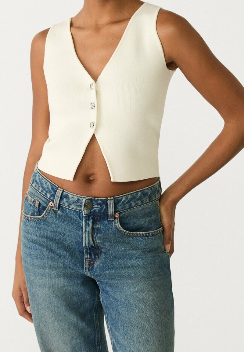 Kvinna i en ärmlös krämfärgad knäppt crop top och högmidjade blå denimjeans med en hand på höften.