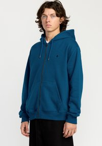 Sudadera con cremallera en color azul verdoso, hecha de una tela suave. Cuenta con un bolsillo frontal tipo canguro, capucha ajustable con cordones y puños y dobladillo acanalados.