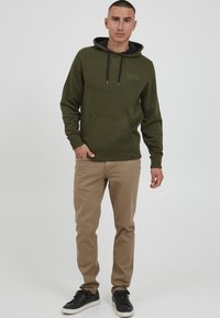 Olivgrüner Pullover-Hoodie mit einer Fronttasche, verstellbarem Kordelzug und gerippten Bündchen, kombiniert mit beigen Hosen und schwarzen Turnschuhen.