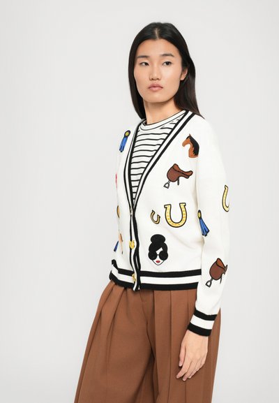 Cardigan bianco con rifiniture nere, caratterizzato da motivi equestri ricamati colorati come cavalli, selle e nastri. Abbinato a una maglietta a righe.