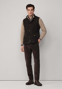Gilet trapuntato nero sopra un maglione beige, pantaloni marrone scuro e scarpe marroni. Caratterizzato da accessori in oro e un design con colletto. Stile minimalista.