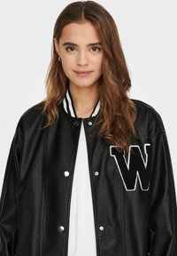 Jachetă de varsity din piele neagră, cu nasturi tip snap, guler cu dungi albe și un patch mare cu litera alb�ă "W" pe pieptul stâng.