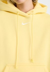 Žuta dukserica s kapuljačom od mekog materijala, s bijelim Nike logom, vezicom za kapuljaču i kengur džepom na prednjoj strani.