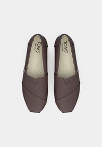 Ζεύγος σκούρων καφέ πανιών TOMS, τύπου slip-on, που απεικονίζονται από ψηλά σε λευκό φόντο.