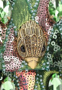 Pezzo decorativo in bronzo con intricati motivi a piuma e accenti neri, circondato da paillettes e perline verdi su un tessuto verde testurizzato.