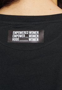 Camisa de tela negra con una etiqueta que presenta el texto "MUJERES EMPODERADAS", "EMPODER___MUJERES" y "HUGO___MUJERES" en una fuente blanca en negrita.