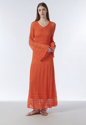 Young woman wearing a long orange crocheted dress with bell sleeves and beige sandals, standing against a plain light background.