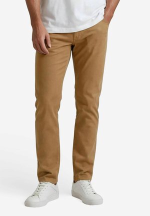 Homme portant un pantalon beige ajusté, des baskets blanches et une chemise blanche, debout avec une main dans la poche devant un fond uni.