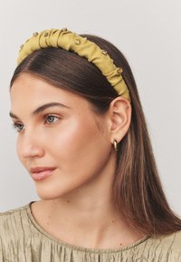 Mustard yellow fabric headband with gathered texture, embellished with gold-tone rhinestones along the top edge. Smooth finish and plush design.
