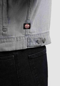 Veste en denim gris clair avec un patch logo Khujo rouge et des pattes à boutons ajustables, portée sur un pantalon noir.