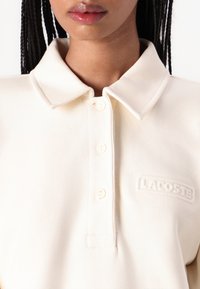 Kremfarget polo-skjorte med strukturert stoff, knappestolpe og brodert "LACOSTE"-logo på venstre bryst. Kort krage.