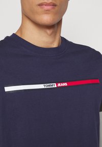 Námořnická modrá bavlněná tričko s kulatým výstřihem a grafickým logem, které má bílé, červené a černé vodorovné pruhy přes hruď.