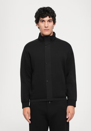 Veste zippée noire avec un col haut, dotée de boutons de fermeture, de poches latérales et d'une texture de tissu lisse. Portée avec un pantalon assorti.