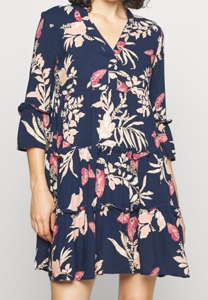 Femme portant une robe bleu marine avec des motifs floraux et papillons roses, avec une jupe à volants et des manches trois-quarts.
