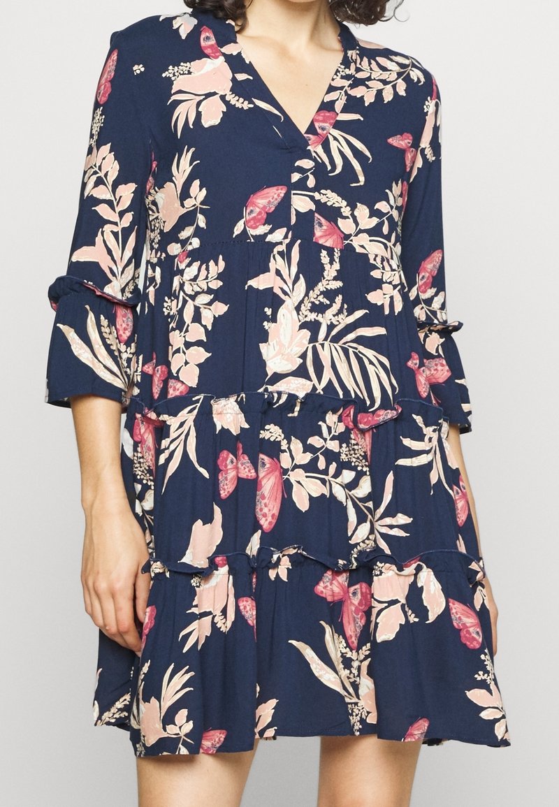 Femme portant une robe bleu marine avec des motifs floraux et papillons roses, avec une jupe à volants et des manches trois-quarts.