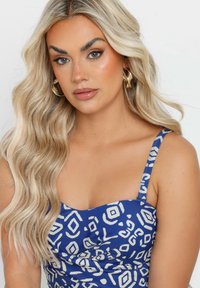 Robe bleue avec des motifs géométriques blancs, dotée d'un corsage ajusté et de fines bretelles. De longs cheveux blonds ondulés et des boucles d'oreilles créoles complètent le look.