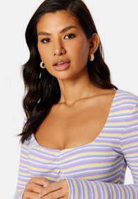 Fioletowa i biała bluzka w paski ze kwadratowym dekoltem, teksturowanym materiałem i długimi rękawami. Złote kolczyki w kształcie kółek uzupełniają całość.