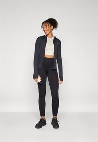 Fekete zipzáras dzseki, krémszínű cropped top, magas derékú fekete leggings és fekete sportcipő. A modell vízpalackot tart, sima háttér előtt áll.
