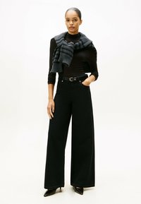 Zwarte wijde broek met hoge taille, gecombineerd met een zwarte coltrui. Een grijs gestreepte gebreide sjaal hangt over de schouders. Glanzende zwarte hakken.
