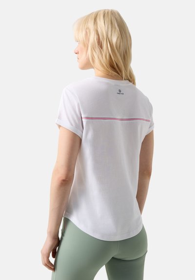 Frau mit schulterlangen blonden Haaren, die ein weißes T-Shirt mit pinken Streifen und salbeigrüne Leggings trägt, von hinten vor weißem Hintergrund.