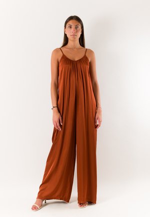 Donna in piedi rivolta in avanti che indossa una tuta larga senza maniche color ruggine con spalline sottili e tacchi aperti.