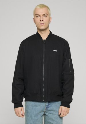 Joven con cabello rubio corto, vestido con una chaqueta negra con cremallera y el texto "UNFAIR" y jeans azul claro, posando frente a un fondo gris.