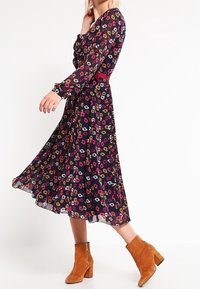 Langärmlig, marineblaues Kleid mit Blumenmuster, das mit kleinen, bunten Blumen, einer taillierten Taille und einem ausgestellten Rock ausgestattet ist; kombiniert mit beige-farbenen Stiefeletten.