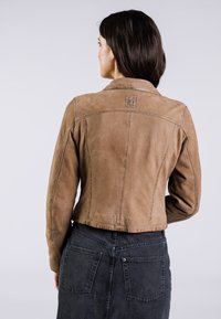 Veste en daim marron au design ajusté, avec des coutures visibles et un petit logo dans le haut du dos. Portée sur une jupe en denim foncé.
