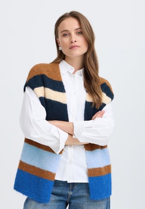 Frau mit langen Haaren, die ein weißes Hemd und eine braune, blaue, cremefarbene und marinegestreifte ärmellose Strickweste trägt, die Arme verschränkt.