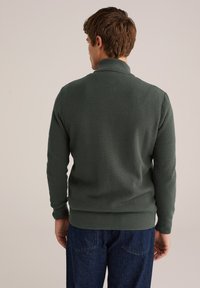 Zeleno, teksturirano pulover s visokim ovratnikom, rebrastimi manšetami in robom. Nosi se preko temnih jeans hlač. Pogled s hrbta.