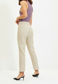 Vrouw in beige high-waisted tapered broek, paarse mouwloze top en zwarte spitse slingback hakken, staand tegen een witte achtergrond.